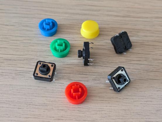 A photo of some small tactile buttons scattered on a desk. There are also brightly coloured round tops to the buttons that clearly just snap on to the top of the buttons to give them an easier to press surface.