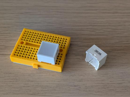 A photo of two small LED lights. They are in a 1.5cm square housing and will light up blue. One is plugged into a small electronic breadboard, the other is on its side and shows the text C00106.