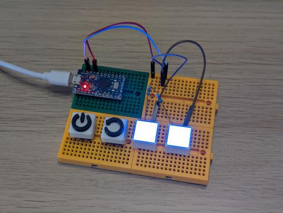 A photo of some electronics on a breadboard. A microcontroller is feeding 5 volts and ground to two square LEDs, which are lit up very brightly blue. Next to them are two buttons marked power and reset but these aren't hooked up to anything.
