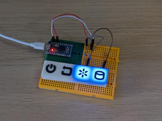 A photo of some electronics on a breadboard. A microcontroller is feeding power to two LED lights underneath a 3D printed panel. That panel features power and reset buttons, and sun and cylinder symbols that the lights are shining through.