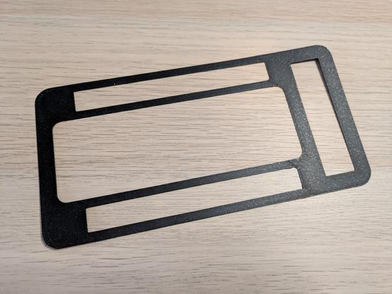 A photo of a very thin, flat 3D print of a PC case front. It's rectangular with large open spaces in the middle. It's black in colour, with both a matte finish and sparkly bits.