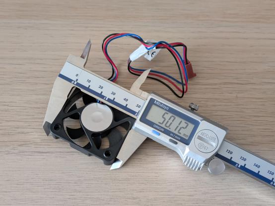 A photo of a small computer fan, with its width being measured at 50.12mm by a set of digital calipers.