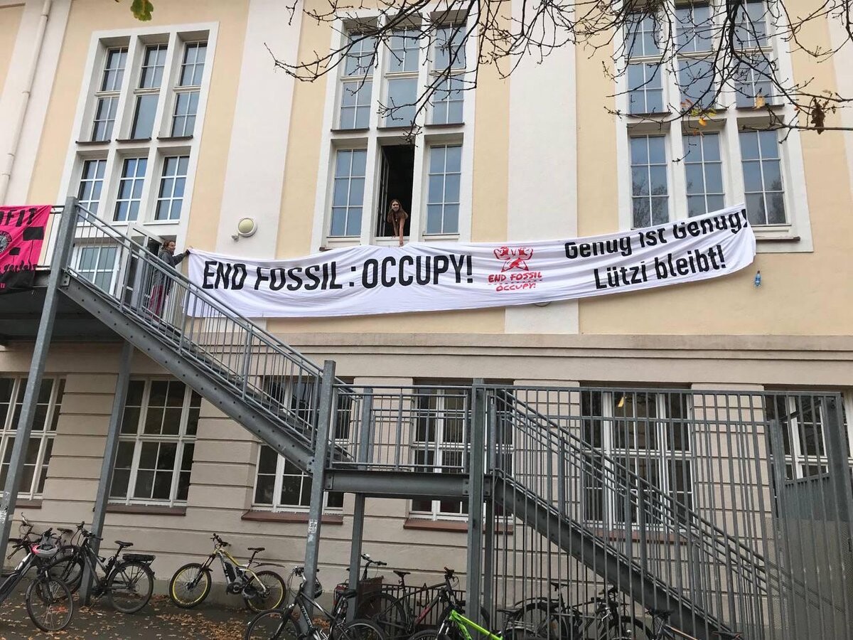 Ein weißes Banner mit der Aufschrift "End Fossil:Occupy-Genug ist genug! Lützi Bleibt!" Vor den Fenstern eines gelben Schulgebäudes. Davor eine silbern-matt glänzende Fluchttreppe woran Fahrräder geparkt sind