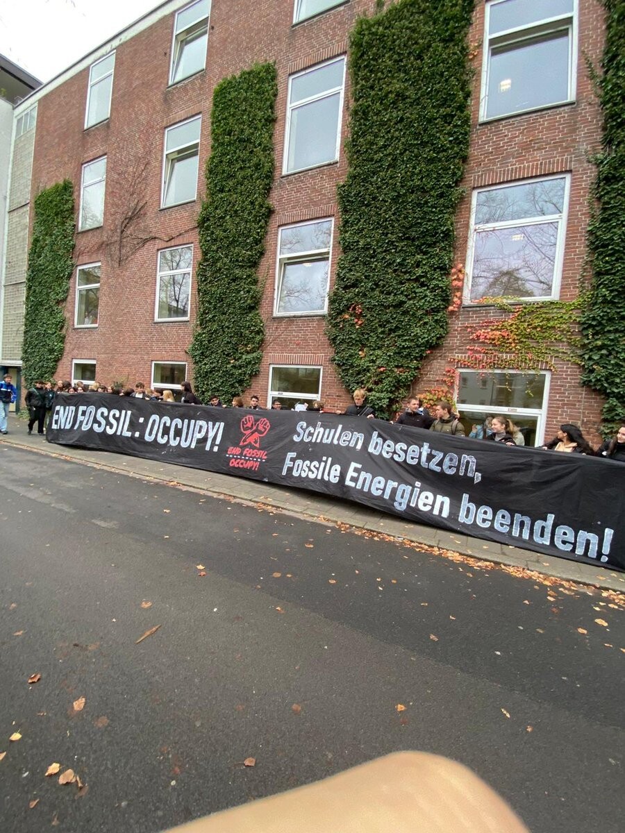Ein schwarzes Banner mit der Aufschrift "End Fossil:Occupy -Schule besetzen, Fossile Energien beenden" vor einem roten Backsteingebäude. Hinter dem Banner stehen ca ein Dutzend Aktivist*innen