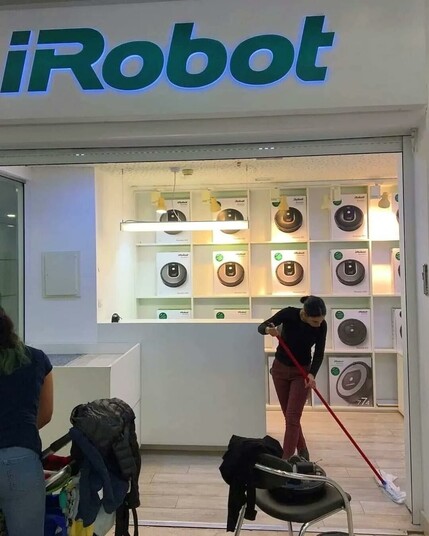 The store front of a small iRobot shop, possibly in a mall. Many vacuum robots of their brand are prominently displayed on the back wall. In front of them, someone is cleaning the floor with a broom.