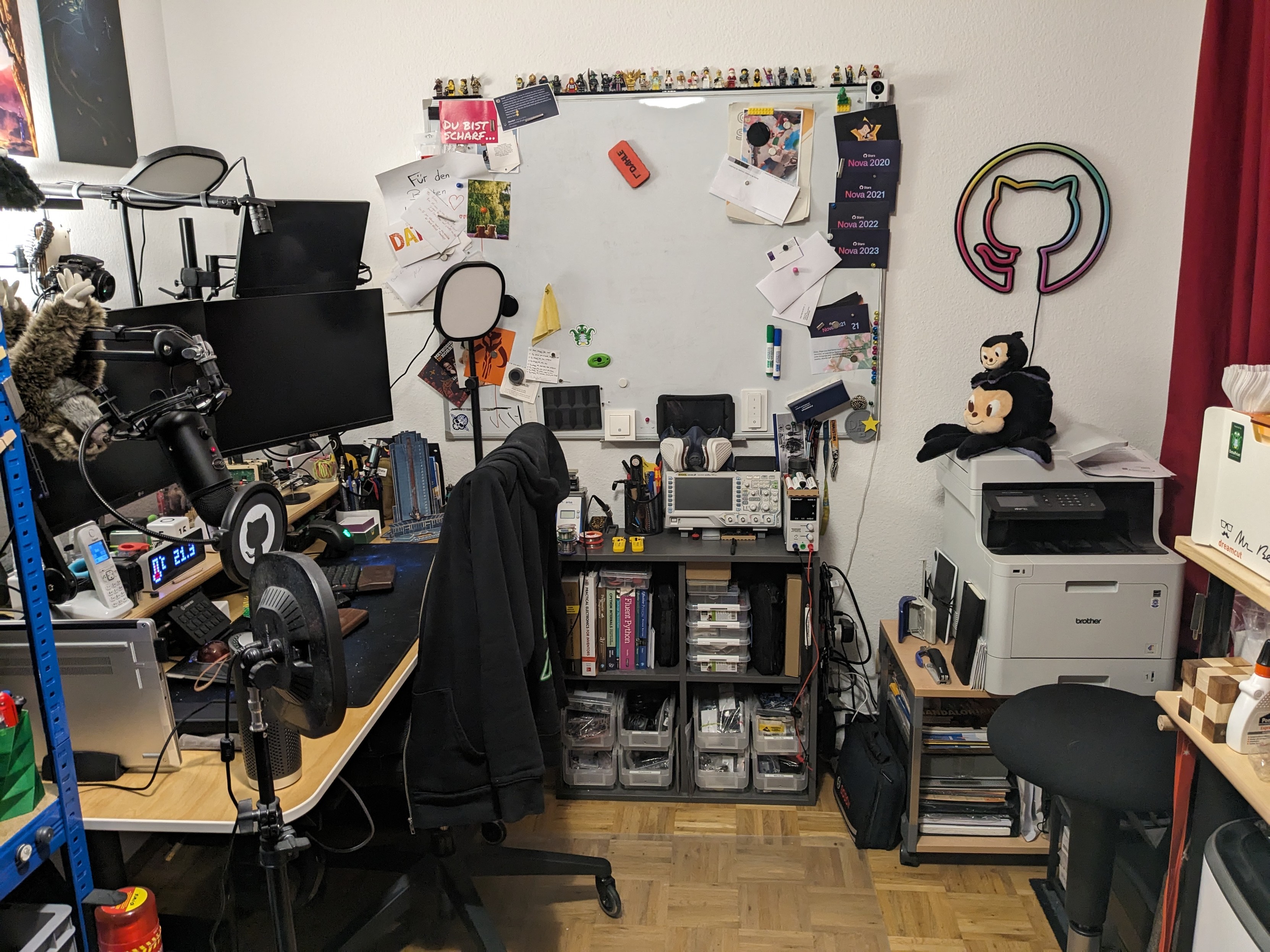 View towards the back of the office. Desk to the left, a big whiteboard and some shelves filled with electronics directly forward and a paper printer and a table with a laser cutter to the right.