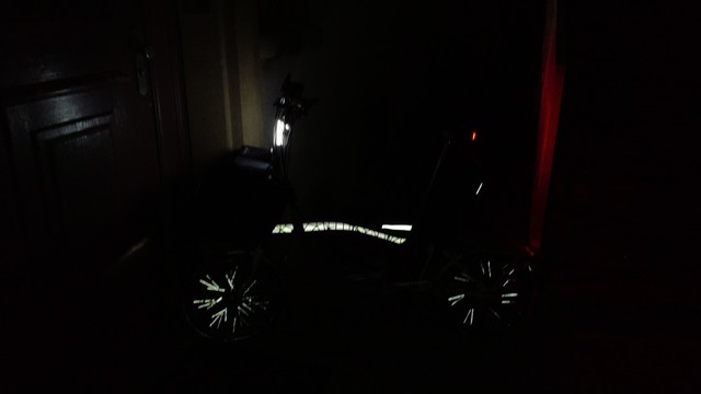 A Brompton folding bicycle in the dark leaning on some door frame, visible from the side. The frame's main tube, some of the spokes, a small part of the saddle pouch and a line on a frame bag are reflecting light from the view point.

Additionally two very bright battery lights are on: A vertical, very bright white one on the handlebar, and a red, less visible one lighting up a fence post near the rear of the bicycle.