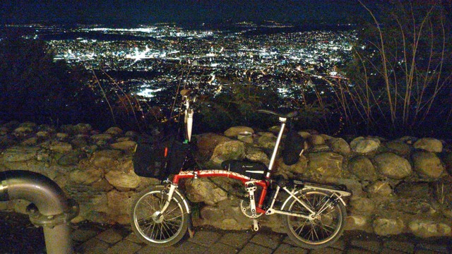 Ein rot-weißes Brompton-Faltrad steht bei Nacht angelehnt an eine kniehohe Mauer aus Wackersteinen. Im Hintergrund Zürich bei Nacht.