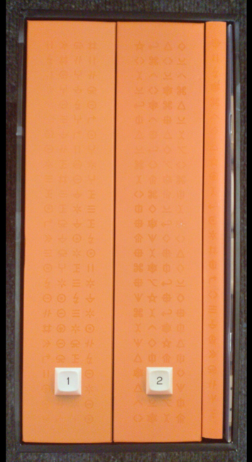 Front view of the Shift Happens slipcase withe two thick and one thinner orange book inside. The two thicker books have beige key caps with legends 1 and 2 as label for volume 1 and 2. There's also a thin, stapled leaflet on the very right end of the slipcase visible.