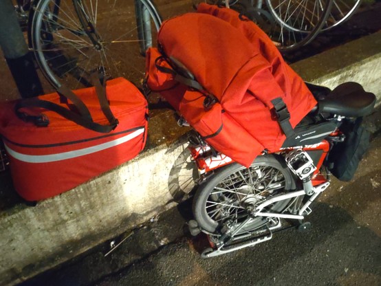 A folded red-white Brompton folding bicycle with white reflective mosaic stickers on the frame and a filled red frontbag mounted. A red, Brompton specific rear rack bag with a white reflective stripe all around it is standing on a low concrete wall left of the bike. Picture shot without flash. Colors are more realistic in this picture.