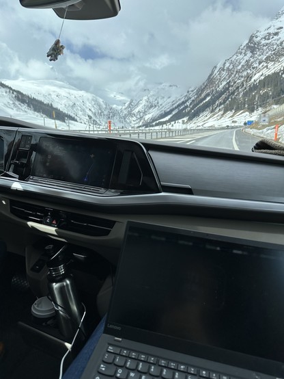 Interior of a car with a dashboard, laptop, and a water bottle, driving on a highway with snow-covered mountains in the background. A small object is hanging from the rearview mirror.