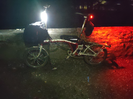 My red-white Brompton folding bicycle leaning on a low wall in the night with rather bright "be seen" lights on the handlebar stem, under the saddle and on the rear end of the rack.