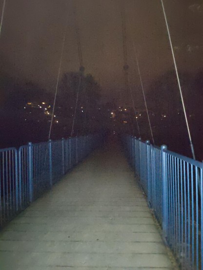 A suspension bridge with blue railings and dusty wooden ground. The night sky is cloudy in sepia tone. Some lights behind trees in the background.