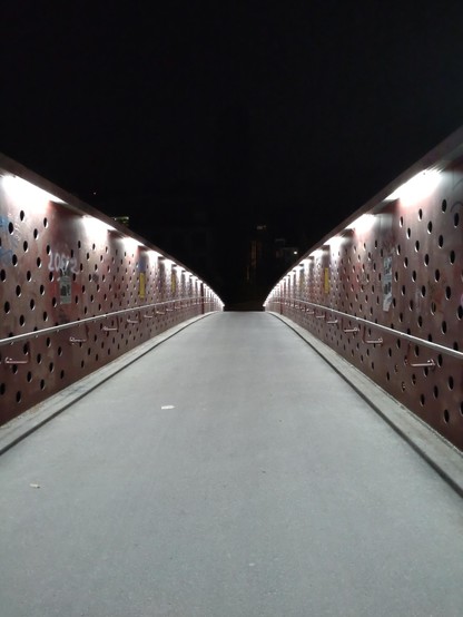 A modern, artsy bridge with red-brown-ish guarding walls with round holes (and quite some graffiti) and neon lights on top shining downwards. Close to the viewpoint the walls are several meters high. But they get much lower towards the end of the bridge. The sky is pitch black.