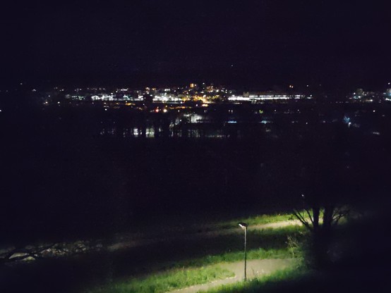 View from a hillslope down into a rather flat valley. Two lighted streets with meadows around them in the front, city lights in the background. In between a dark river which mirrors some of the city lights.