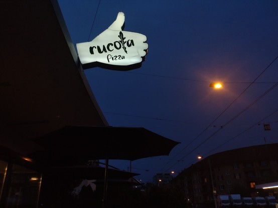 A lighted restaurant sign in form of a thumbs up gesture with the words "rucola" and "Pizza" in it. A dim amber streetlight in the dark blue evening sky.