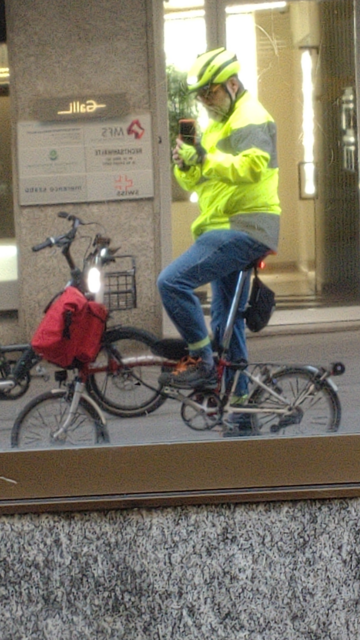 Me (jeans, yellow hi-viz jacket and bicycle helmet) on my red-white Brompton (battery lights on) with a red frontbag reflecting myself in a shop window coated with golden foil.