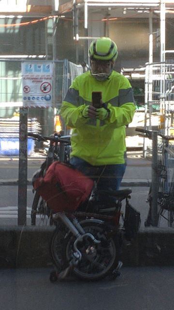 Me (jeans, yellow hi-viz jacket and bicycle helmet) standing in front of my folded red-white Brompton with a red frontbag still mounted; reflecting myself in a foil coated shop window with quite some scratches. (Picture before doing groceries.)