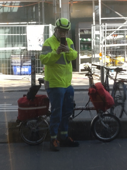 Me (jeans, yellow hi-viz jacket and bicycle helmet) standing in front of my unfolded red-white Brompton with a red frontbag and a rather full red-white rear rack top bag; reflecting myself in a foil coated shop window. (Picture after doing groceries.)