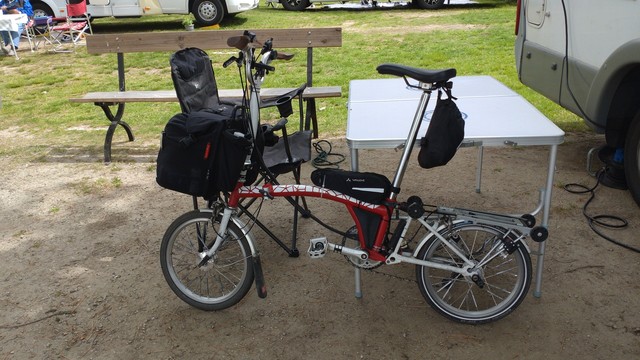 A red-white Brompton with a black front bag leaning on a camping table, ready for riding.