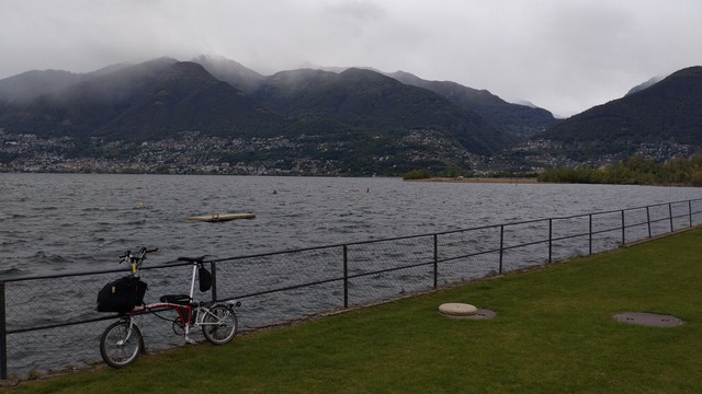 Brompton leaning on a guarding rail at Magadino grass "beach". On the other side of the lake dark green mountains with their tops in clouds and many villages on their lower hillsides. (What's visible is the region between Locarno and Tenero with the one valley cut being the Val Verzasca.)