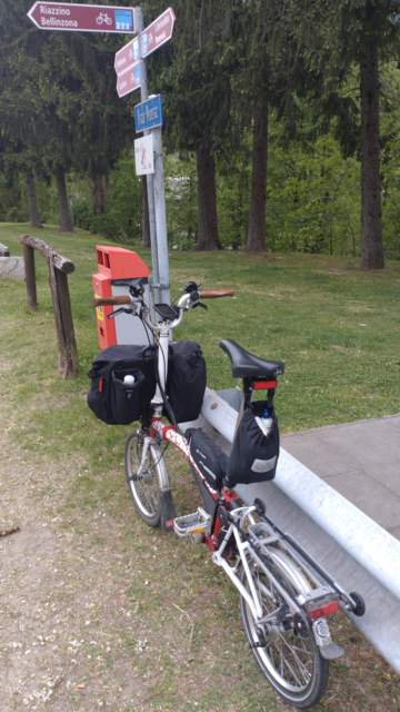 A red-white Brompton leaning on a guarding rail in front of a bicycle path directions sign with 311 and 31 both leading to Bellinzona. This crossing of the two bicycle paths in Arbedo was my target and turning point.