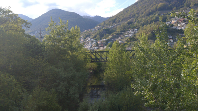 A box-girder bridge reflecting itself in a river. Lots of green in the foreground, a village on a hillside in the background. Dark green mountains in the distance.