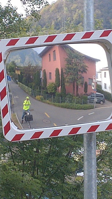 A traffic mirror with red-white frame showing a cyclist in a yellow hi-viz jacket.
