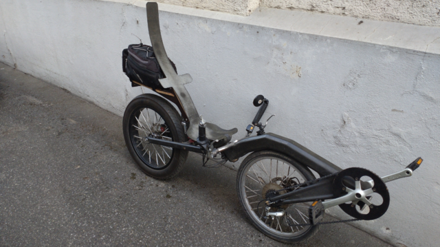 A DIY pivot steering FWD recumbent in the style of a Flevobike made out of carbon frame parts and a rather thick and wide rear wheel.
