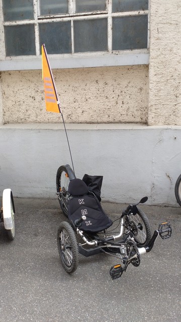 Schwarzes KMX-Liege-Trike mit großem, orangefarbenem Sicherheitswimpel von schräg vorne.