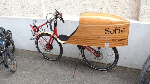 "Sofie" a red, front wheel driven short wheelbase recumbent with an aerodynamic wooden box in the back which also provides the seat. It has lettering on the box that says "Sofie made by holzwurm 2024".

A visitor's bicycle.