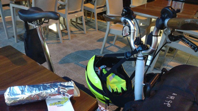 One end of a garden restaurant table with a Falafel Dürüm wrapped in aluminum foil. Besides the table, there's a half-folded Brompton standing with a yellow helmet hanging on the handlebar.