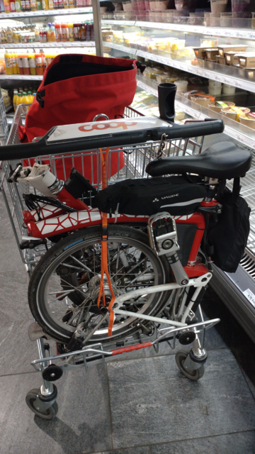 A folded red-white Brompton sitting on a swung out platform at the rear end of a shopping cart originally meant to put crates of beverages there. It's saved against falling off with an orange strap between the bag hook of the cart and the trailer hook of the Brompton
