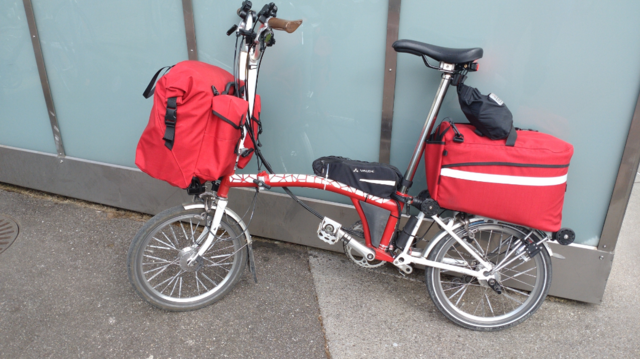 A folded red-white Brompton sitting on a swung out platform at the rear end of a shopping cart originally meant to put crates of beverages there. It's saved against falling off with an orange strap between the bag hook of the cart and the trailer hook of the Brompton