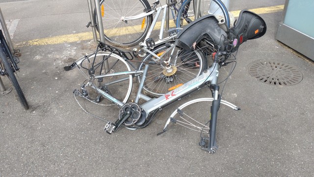 A silver step-through e-bike with mid-engine, but without saddle, wheels and battery, laying on the ground unlocked. Even the battery lock seems ripped out.