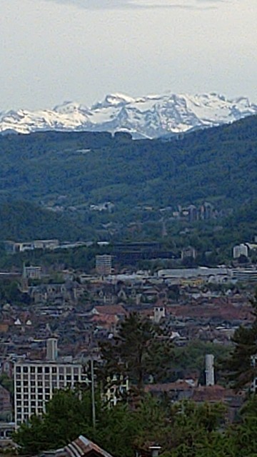 A photograph in portrait orientation showing city roofs and some green trees in the foreground, white mountains in the distance and dark green hills in between.