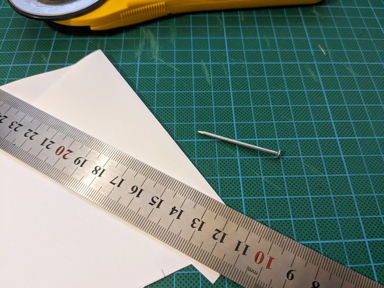 A nail lying on a green scuffed cutting mat. Next to it lies a metal ruler and a just folded sheet of white paper. In the back there is a cutter partially visible.