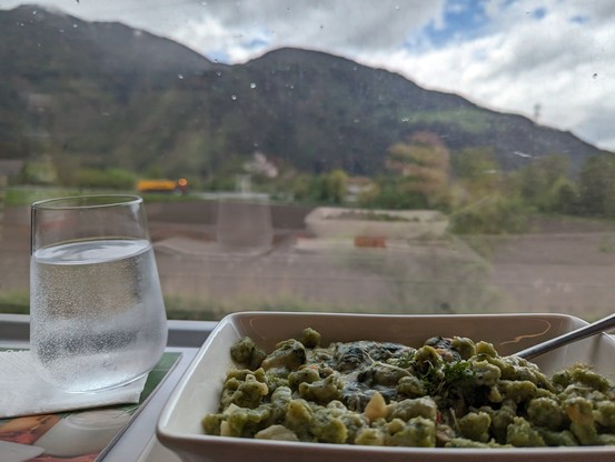 Spinatspätzle und ein Glas Wasser, im Hintergrund unscharf grüne Berge und ein bewölkter Himmel