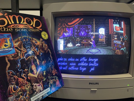 The big box of Simon the Sorcerer showing the gruesome evil wizard, and in the front the teenage wizard Simon together with his dog Chippy and many orcs and goblins. In the background a Philips CRT screen shows the opening scene in the wizard house.