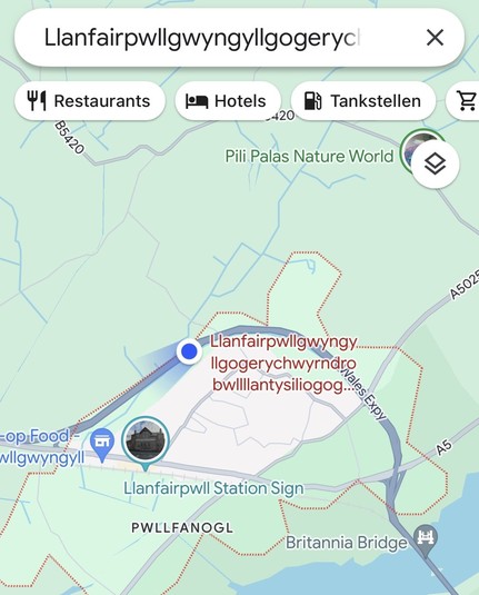 Screenshot Google Maps App, in der die Langform des Namens der walisischen Stadt mit einer Ellipse am Ende abgekürzt werden muss, weil drei Zeilen auf der Karte nicht ausreichen. Auch in der Suchfeld passt der Name nicht komplett rein.

Ein blauer Punkt mit Kompassausrichtungstrichter zeigt dass sich das Gerät auf dem die App läuft gerade auf der Landstraße befindet, die Llanfairpwllgwyngyll außen umfährt.