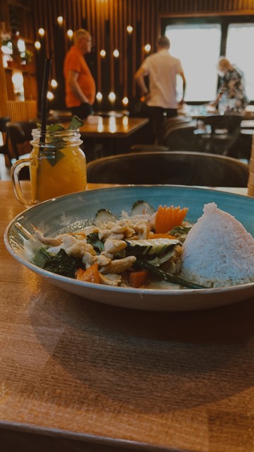 Ein Teller Reis mit Gemüse und Hühnchen, serviert neben einem Einmachglas eines gelben Getränks mit einem Strohhalm, auf einem Holztisch in einem düll beleuchteten Restaurant.
