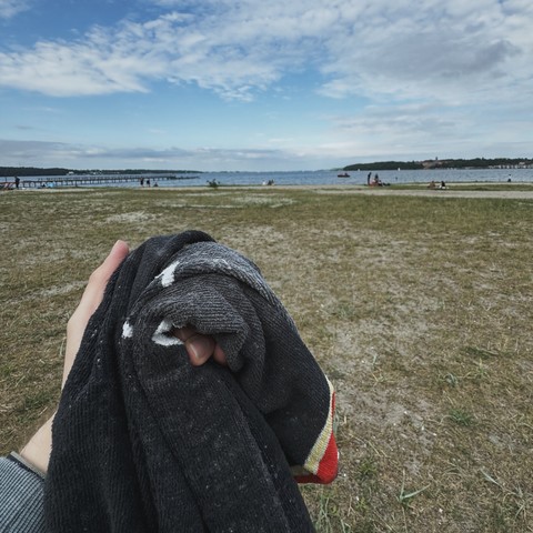 A person holding a rolled-up towel on a grassy beach near the Baltic sea, with a blue sky and scattered clouds in the background.