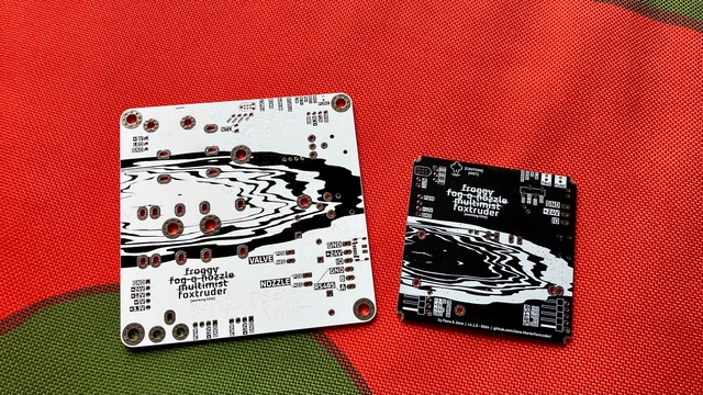Two PCBs next to each other, the left one is white with a black print. It is considerably larger than the right board, which is black with a white print. Both feature an inverse of the same artwork, an abstraction of fog, it almost resembles waves on a round coast. Both boards have plenty port labels and the project title “foxtruder [working title]”. They both rest on some red and green fabric 
