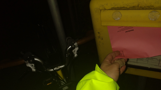Me posting a red voting letter into a rather dirty classic German yellow letterbox. It's dark outside and my Brompton is leaning on a lamp post in the background.