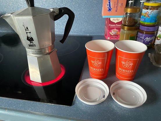 Bialetti auf dem Herd, daneben zwei Coffee-to-Go Becher mit Deckel, orange mit Aufdruck „Werner‘s“

Dahinter Küchenchaos.