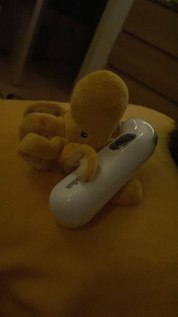Ein gelber Mini-Oktopus sitzt auf einem gelben Kissen und hat ein Ohr-Fieberthermometer in den Armen 