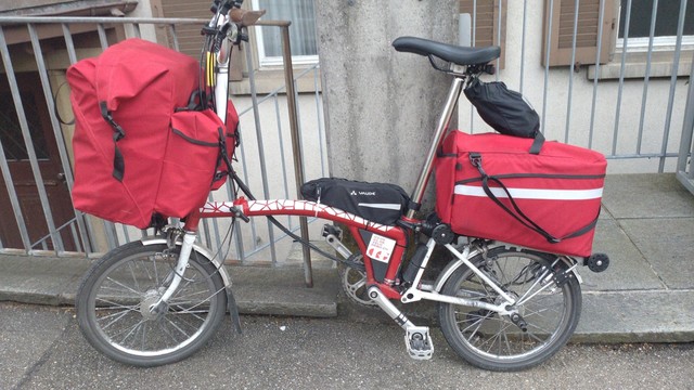 A red-white Brompton is leaning on a concrete post of a guarding rail. It has two red bags mounted, on a the fron and one on the rear rack. Both bags are cram-full. Landscape profile view.