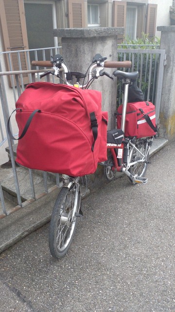 A red-white Brompton is leaning on a concrete post of a guarding rail. It has two red bags mounted, on a the fron and one on the rear rack. Both bags are cram-full. Front quarter portrait view.