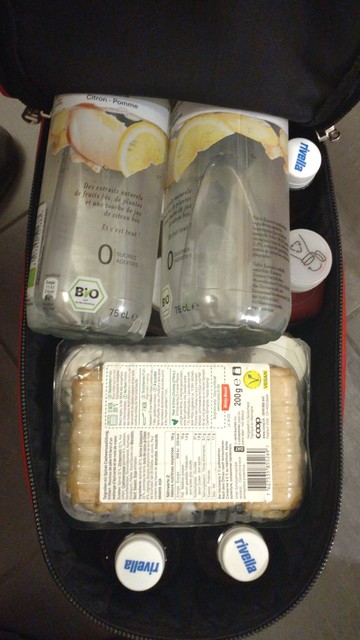 Top-down view on the open ¾ zipper of the rear bag full to the brim with two layers of beverages and one package of vegan spring rolls.