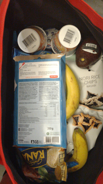 Top-down view on the open roll-top front bag full to the brim. Bananas yogurt, some ready-made vegetarian meal, an avocado, some ready-made pasta and some nori rice chips can be seen.